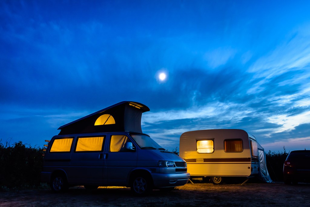A camper van and a caravan illuminated from inside at nightfall with the moon glowing above in a stormy sky.