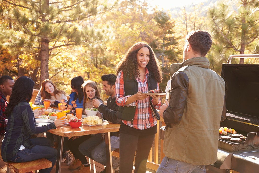 Group of friends eat at table on a fall day while a man at the grill offers a woman a plate.