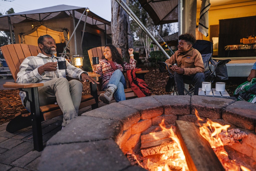 A father and two kids sit in chairs around the campfire at a KOA campground.