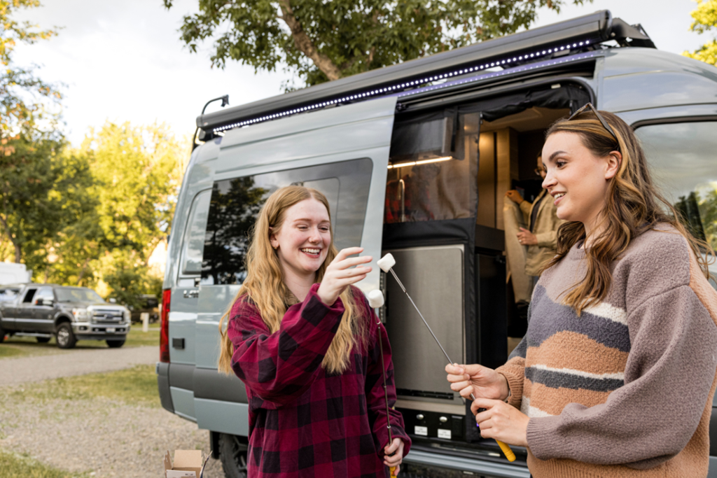 Two women in front of a campervan at Billings KOA Holiday put marshmallows on a stick for s'mores in fall
